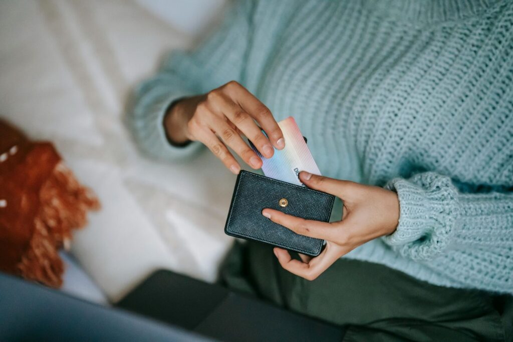 From above crop anonymous female in casual sweater taking credit card from wallet and lying on bed with laptop before making online purchase