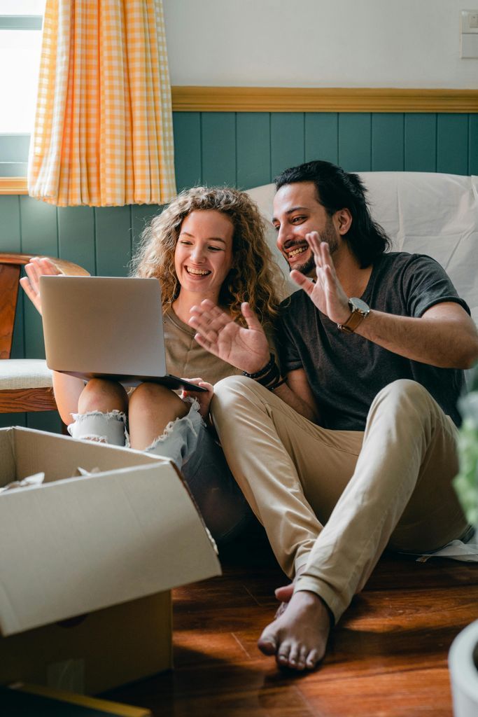 Full body happy smiling diverse couple in casual wear sitting on floor near cardboard boxes in new apartment and chatting via netbook