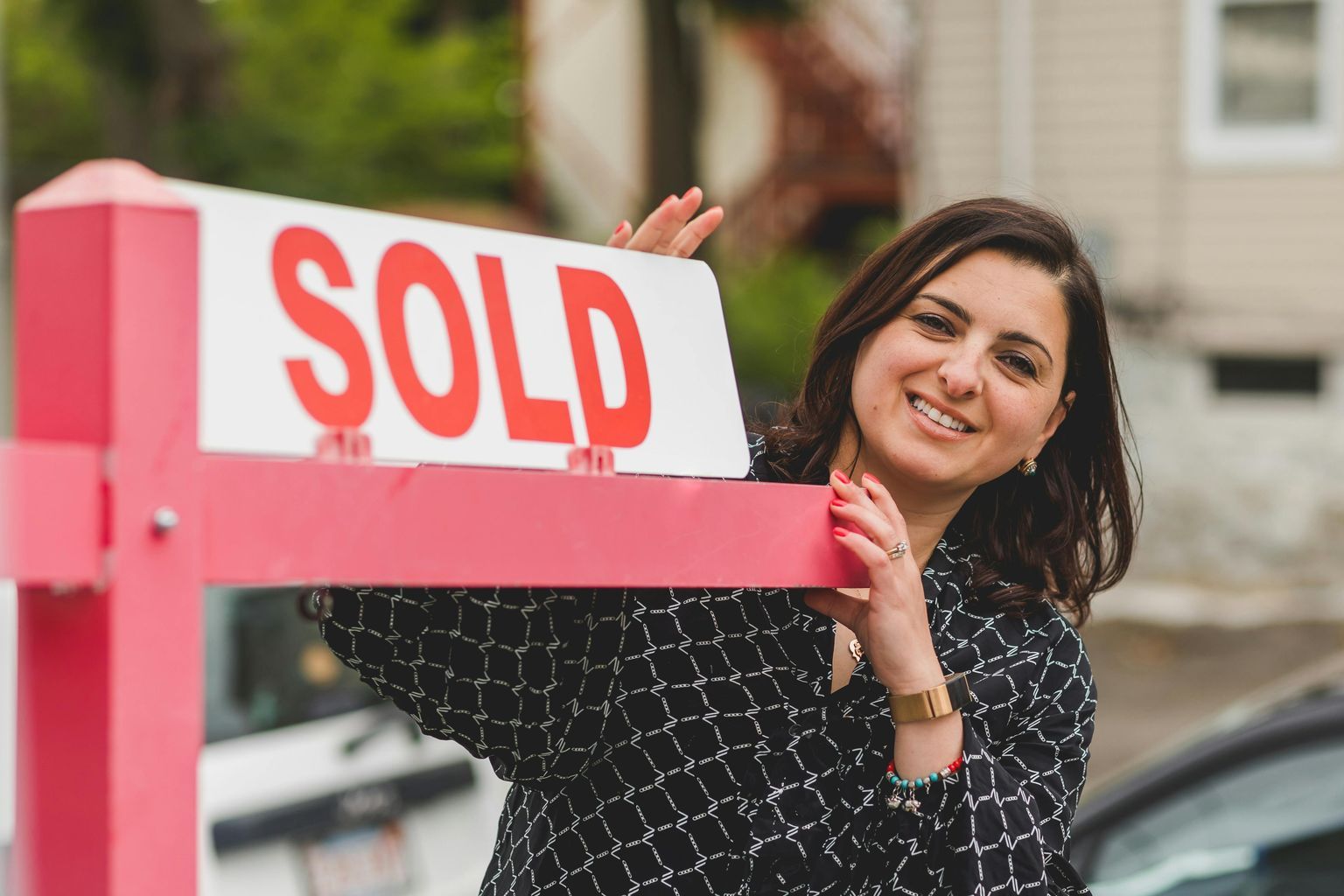 Real Estate Agent Smiling in Black Printed Blouse Holding a Sold Signage Metal Post Outside a House