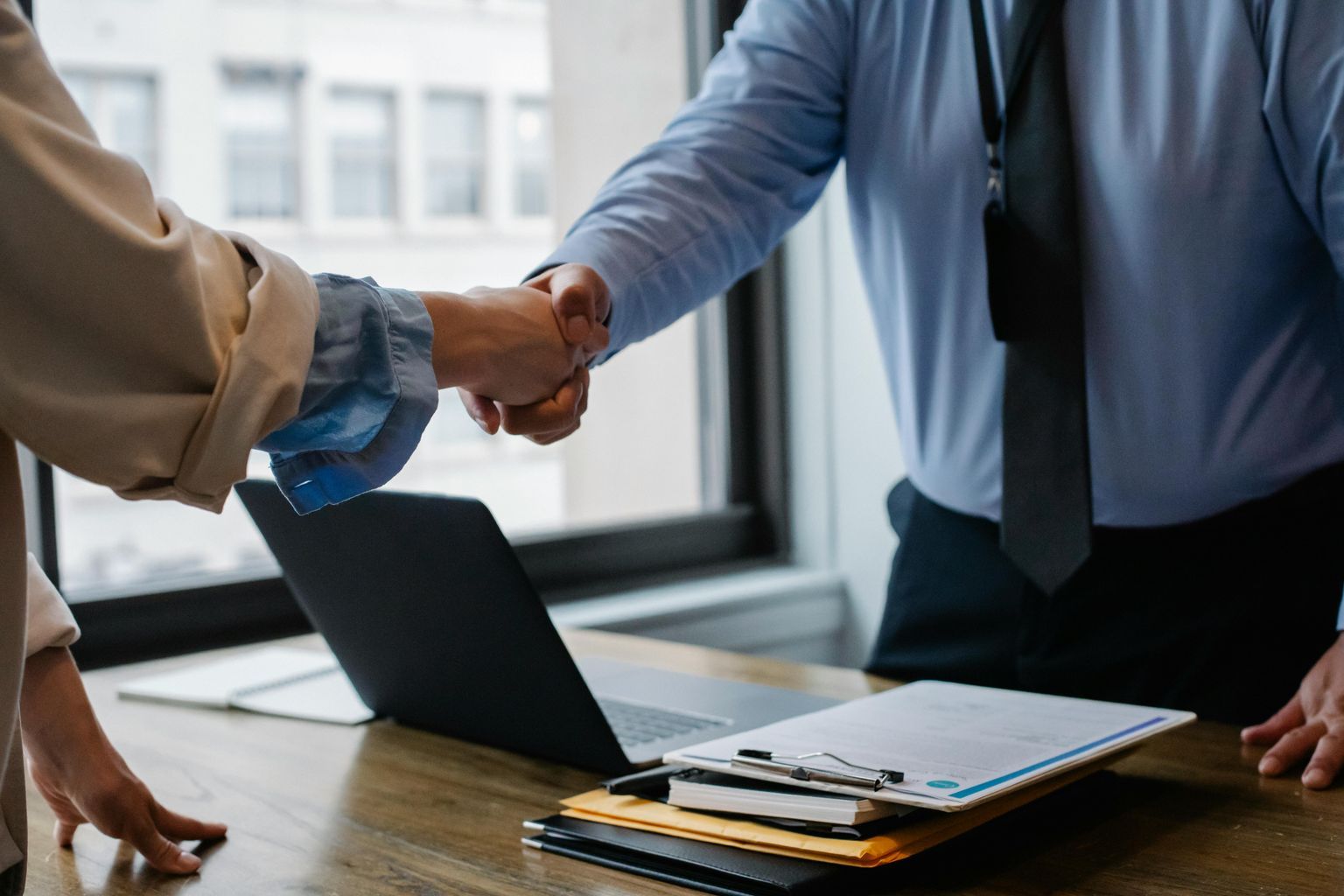 Crop unrecognizable coworkers in formal wear standing at table with laptop and documents while greeting each other before meeting